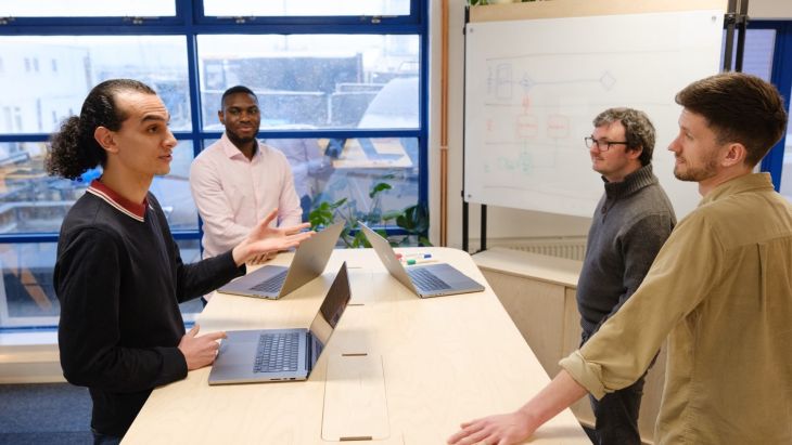 Four men are stood around a table with laptops. One is speaking, indicated by his arm gesture and the other three are looking at him and listening.