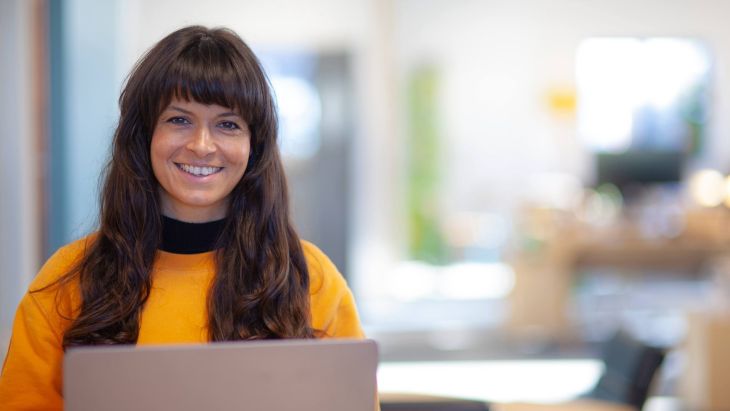 Bianca smiling at her laptop wearing an orange jumper, the studio is blurred in the background