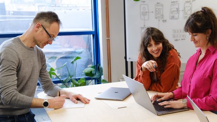 The design team are looking at a laptop screen together, with a white board behind them and James is taking notes on a piece of paper