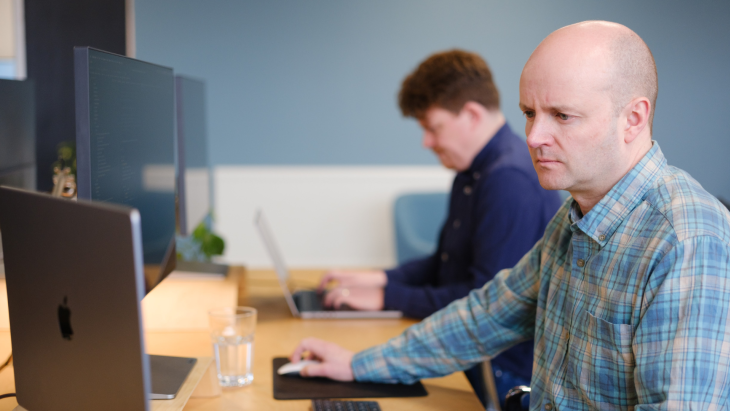 Nick is wearing a checked shirt and working on his laptop, Alistair is working next to him in a dark blue shirt
