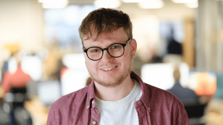Headshot of Rhys, smiling wearing a white t-shirt and pink overshirt.